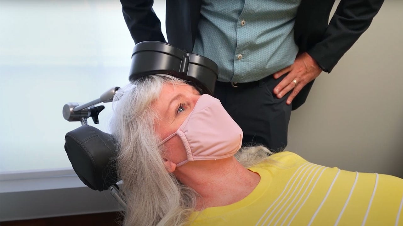 Doctor monitors woman in face mask getting treatment through an electromagnetic coil placed on her head.