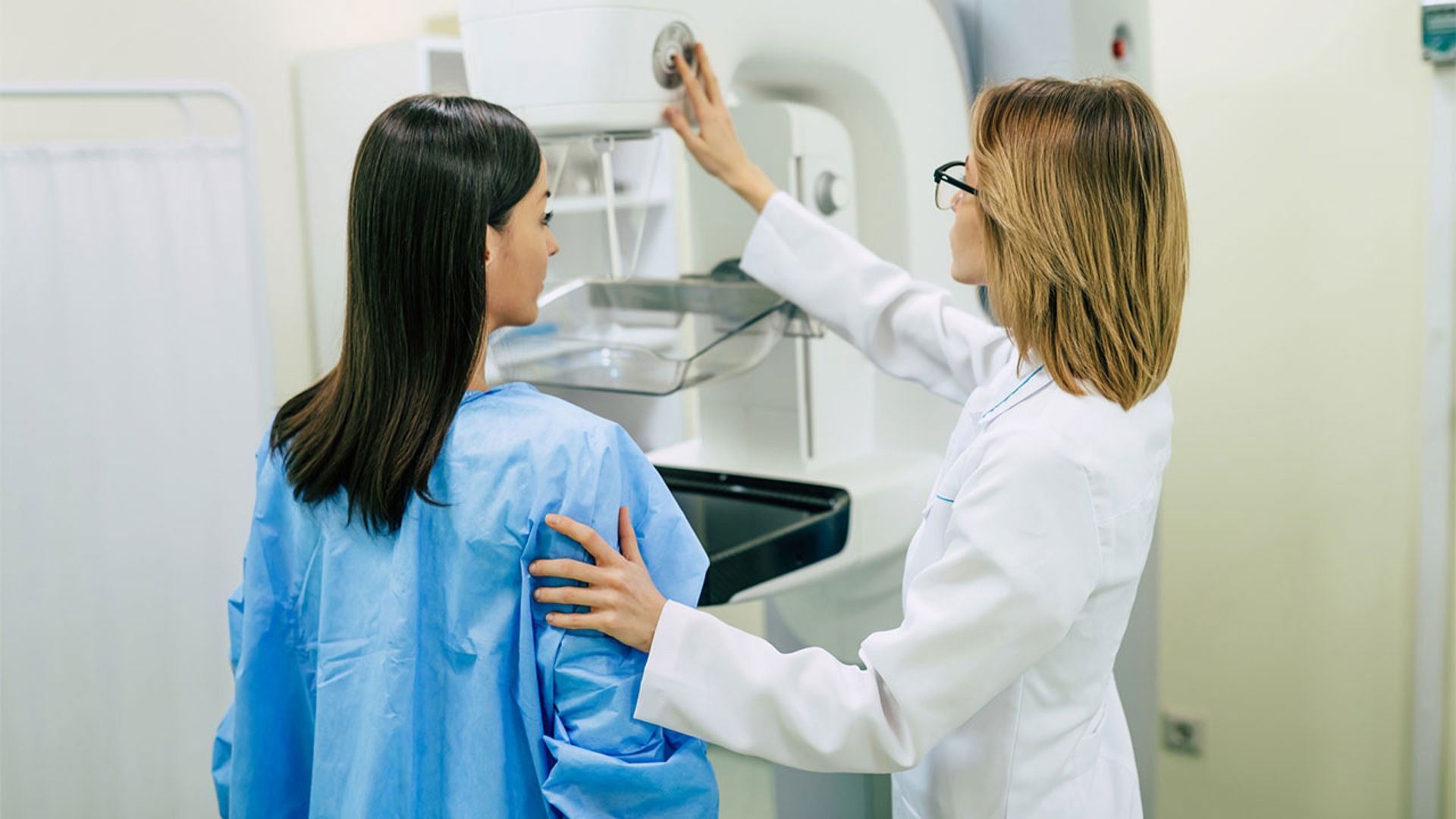 woman in hospital gown looks at medical provider who is adjusting a mammogram machine