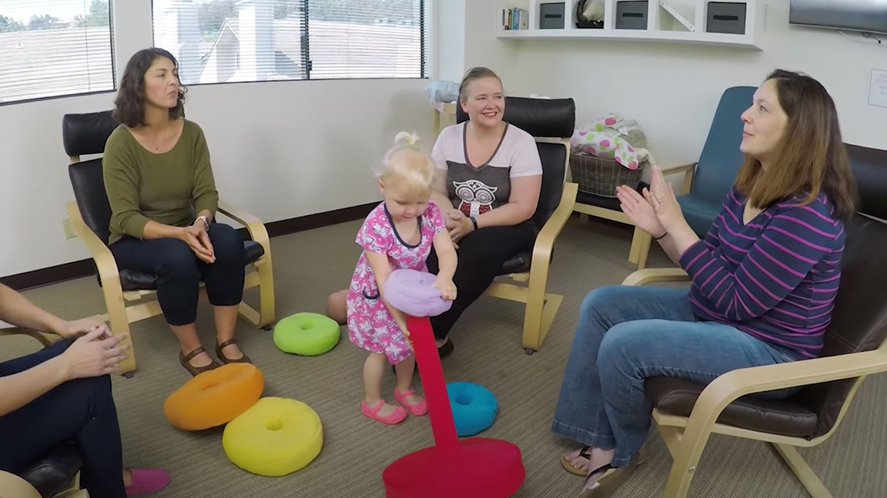 four woman sit around in a room talk while young girl plays with toys in the middle  